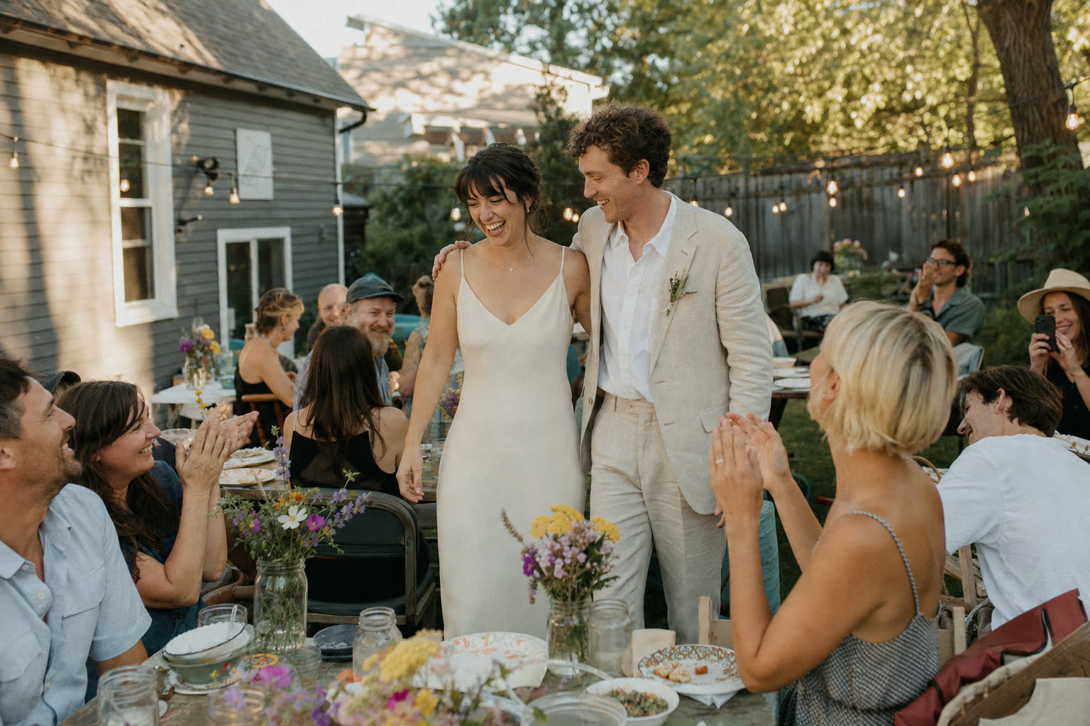 A couple laughing with guests seated around a long table at a backyard wedding in warm evening light